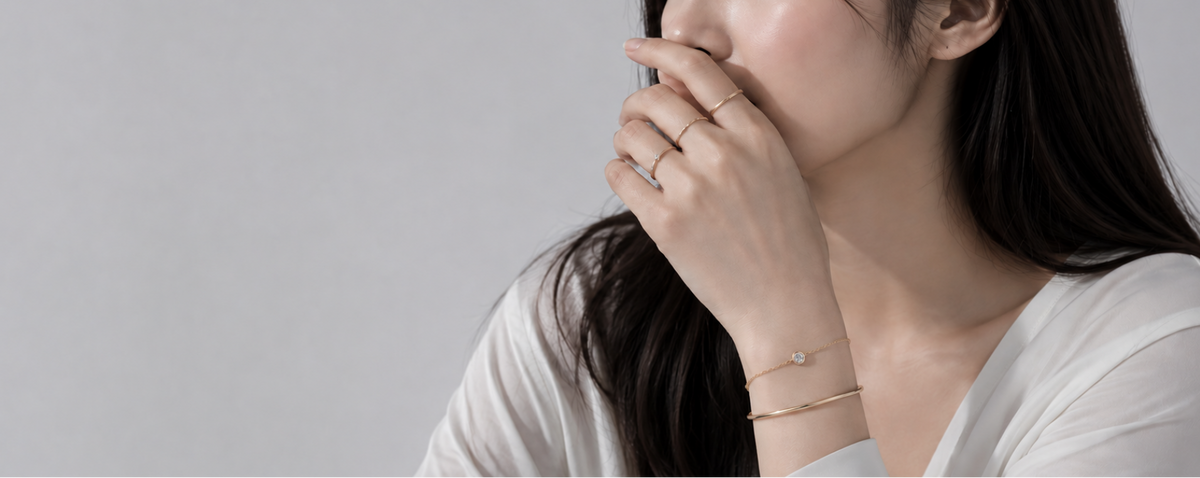 A woman in a white top wearing delicate gold jewelry, including a thin gold ring on her finger and a fine gold chain bracelet on her wrist, against a plain light gray background.