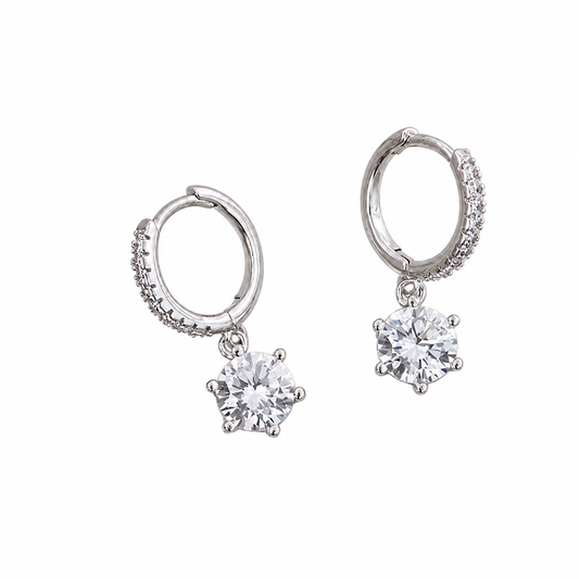 A pair of silver huggie hoop earrings with small pavé accents on the outer edges and a dangling round clear gemstone in a prong setting below each hoop, shown on a plain white background.