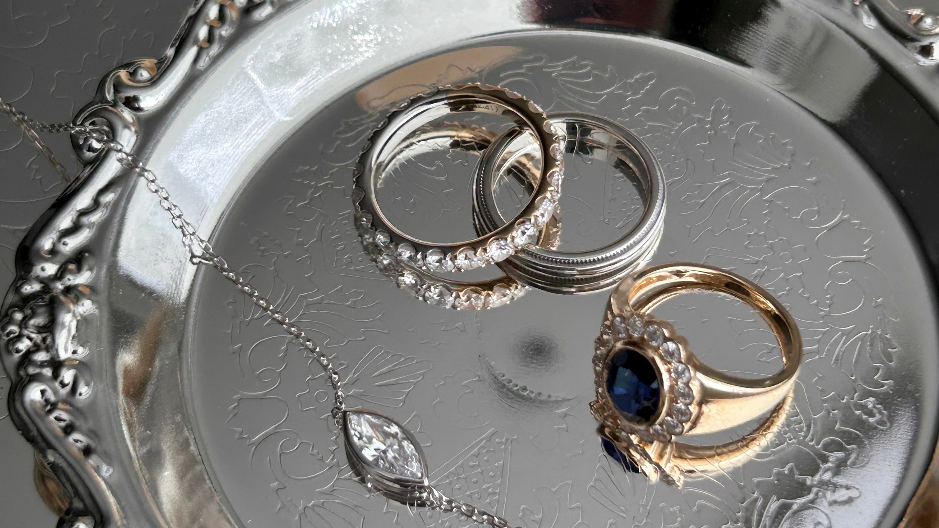 A close-up of jewelry displayed on an ornate silver tray, including two silver rings, a gold ring with a dark blue stone, and a silver pendant necklace, with a perfume bottle blurred in the background. From guesthouse seoul jewellery