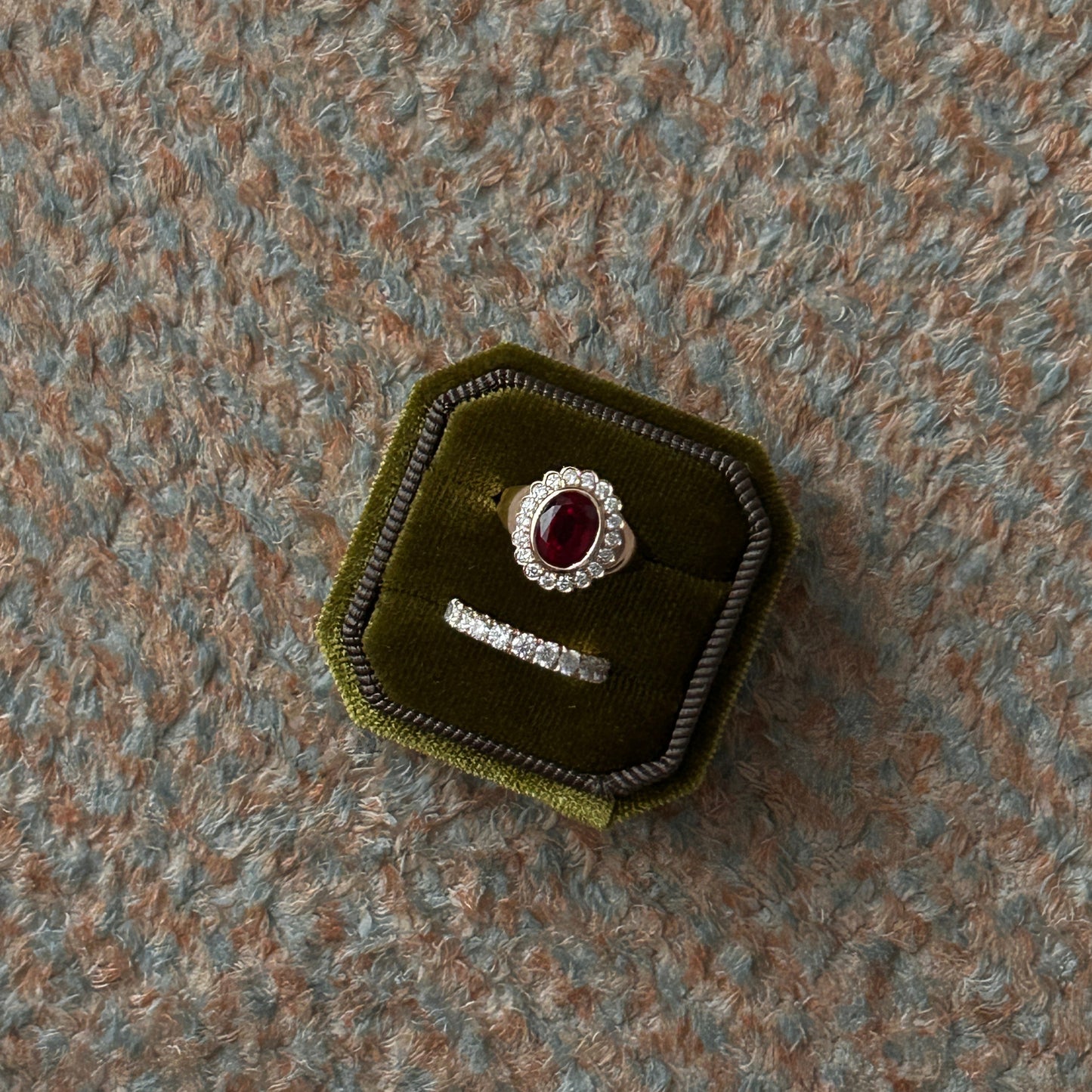 A silver ring set with a round red gemstone and a matching silver band, displayed inside an octagonal dark green velvet jewelry box on a textured gray carpet.
